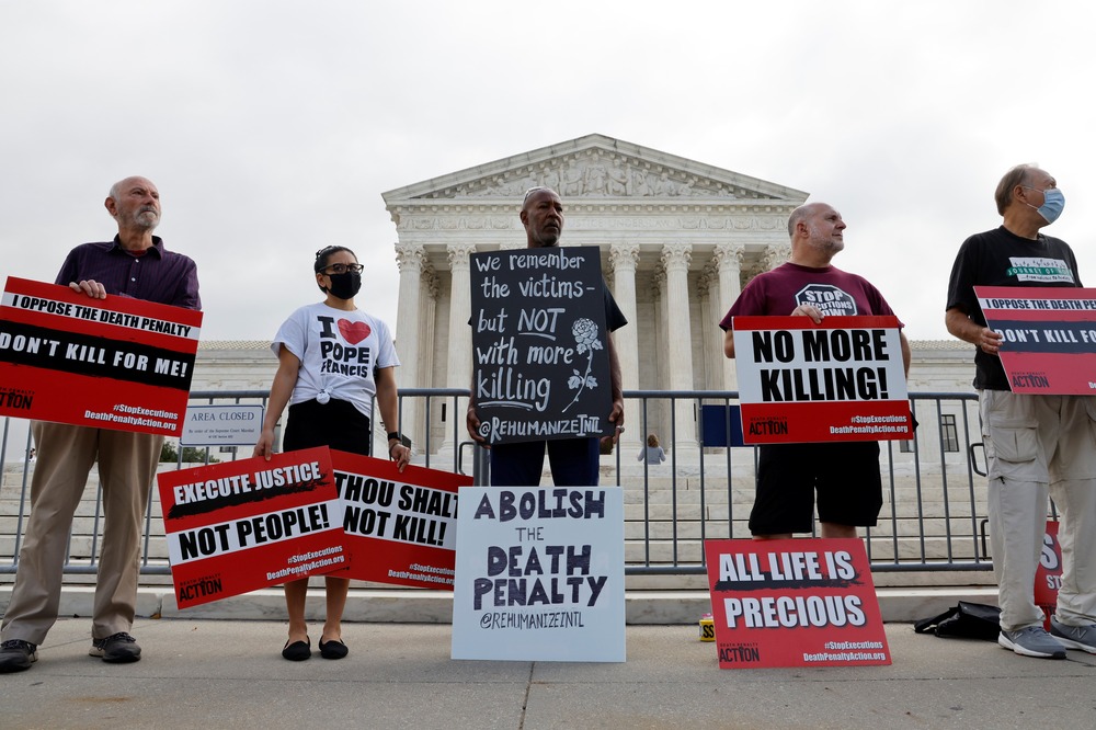 5 people stand in front of Supreme Court building holding signs. 