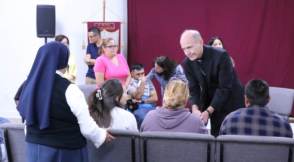 People sit and stand, some crying. The bishop bends down to shake a hand.