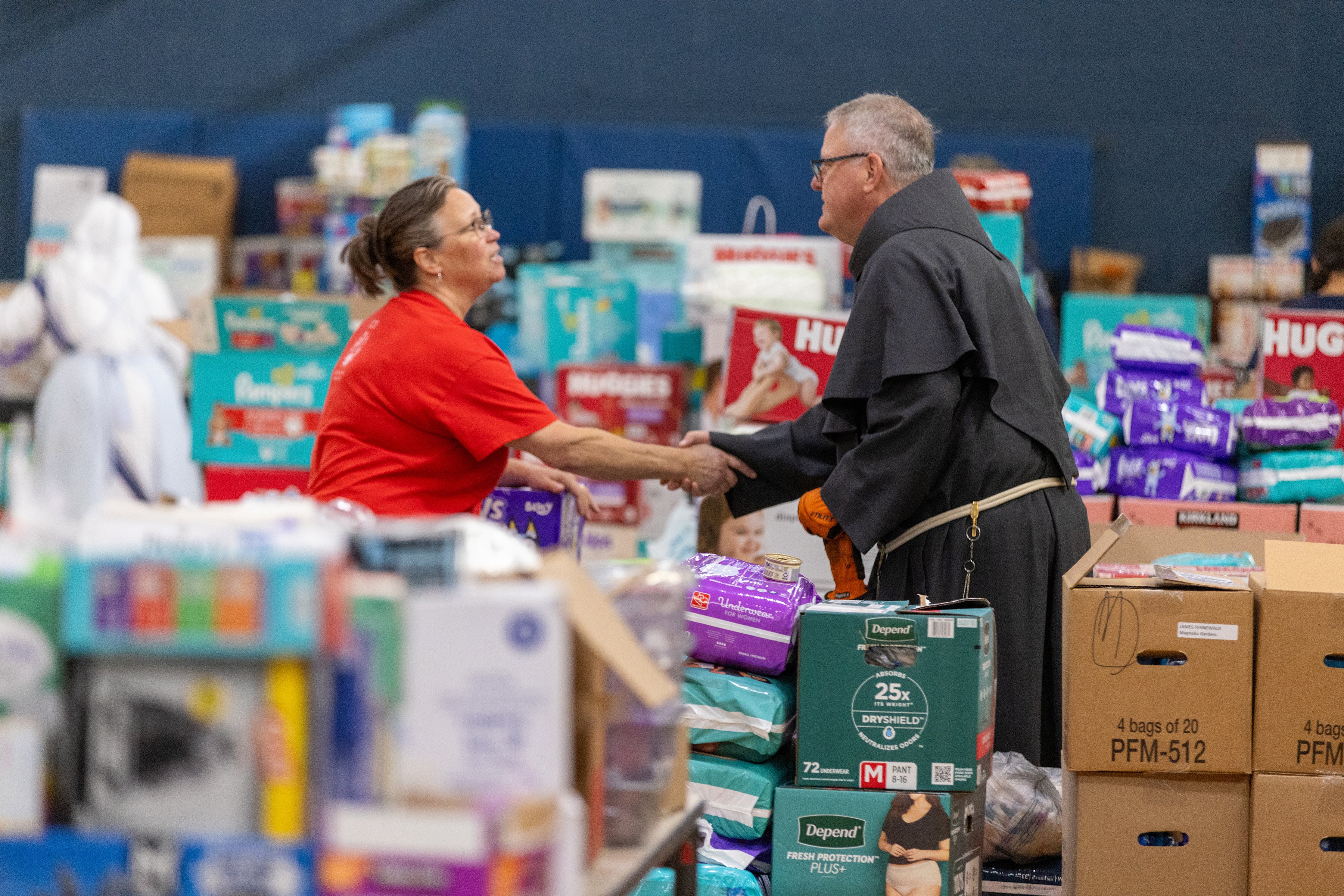 The bishop, wearing habit, shakes hands with a woman. The two stand amid large stockpile of supplies.