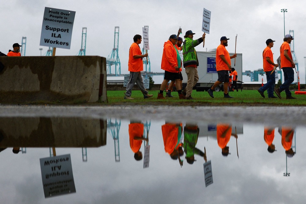 Picket line viewed from side, as workers wearing reflective orange gear and holding signs walk by; they are reflected in a pool of water that occupies the bottom half of the frame. 