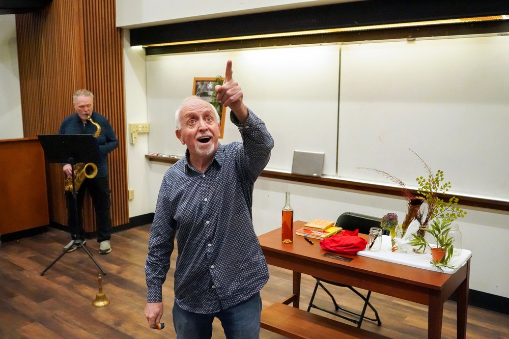 Sandford looks up and out pointing, behind him is classroom whiteboard and Benoit with saxophone.