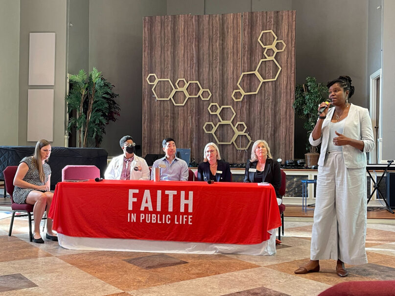 Lewis stands speaking into mic in front of panel with five other people seated. 