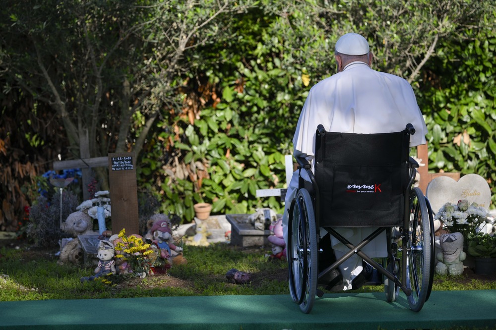 Francis pictured from behind, seated in wheelchair, alone and facing small graves adorned with votives.