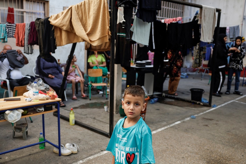 Young boy wearing oversized shirt foregrounded; in background are other people, and a metal frame draped with cloth.