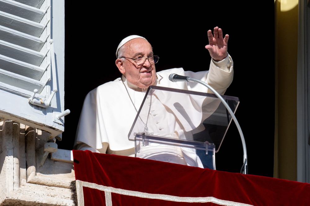 The pope stands at lectern in window and waves to people. 