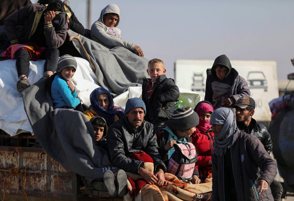 Adults and children, bundled up and carrying items, sit on back of vehicle.