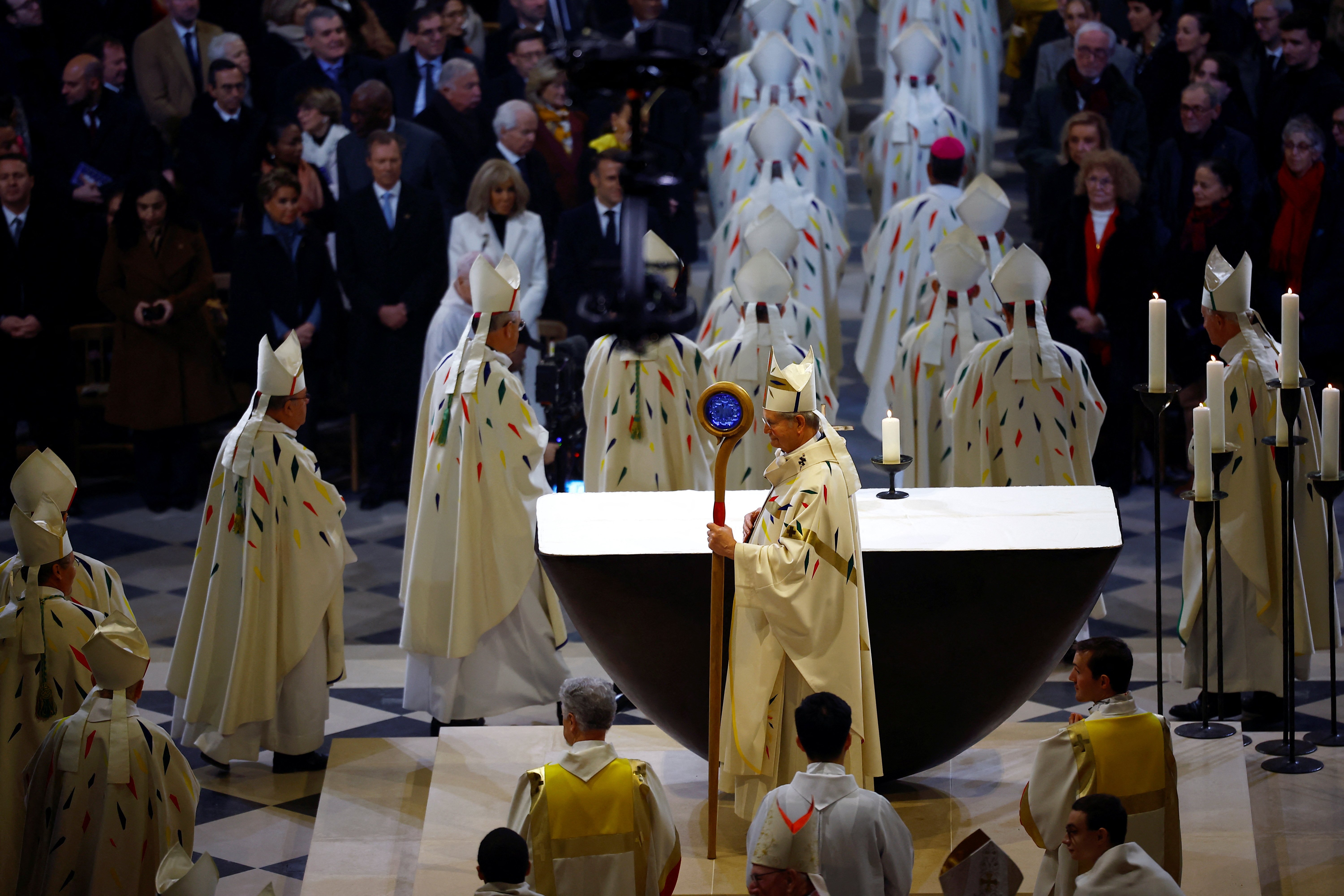 Archbishop, fully vested, pictured in profile holding crozier with massive blue stone; other bishops process, and seated in front of right row are France's presidential couple.