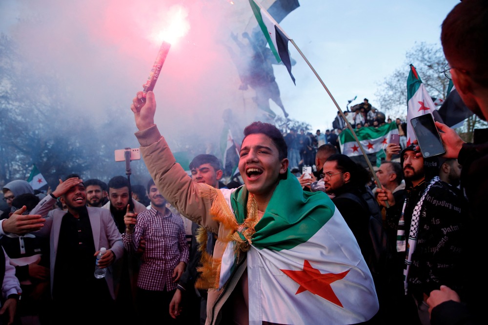 Overjoyed young man wearing Syrian flag, holds flare and smiles; behind him is crowd.