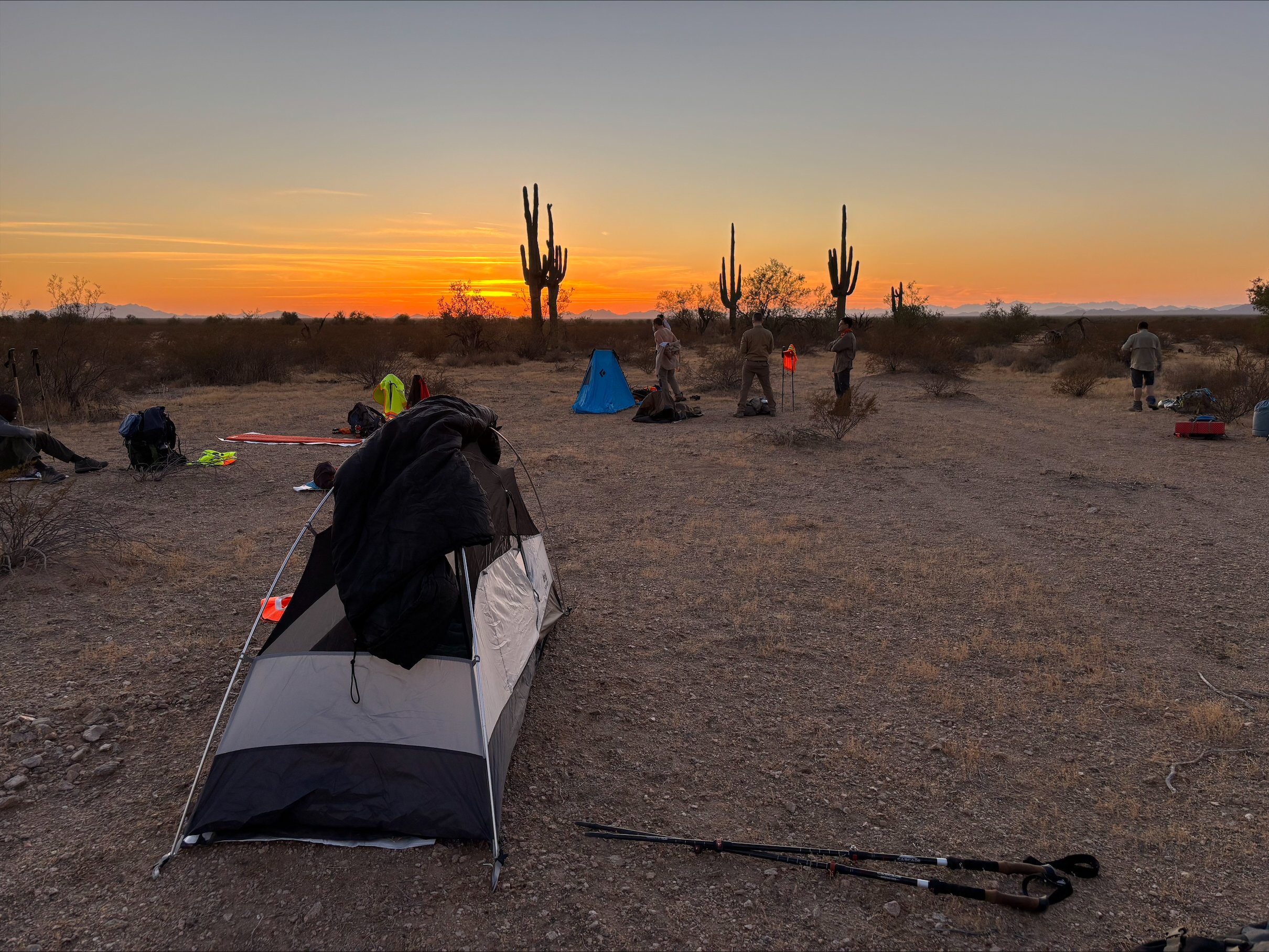 Desert panorama lit with orange hues of sunset, foregrounded is a tent and people sitting and walking.