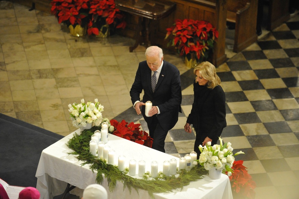 President Biden and First Lady Jill Biden wear black and approach altar adorned with greenery and candles. The president holds large white pillar candle. 