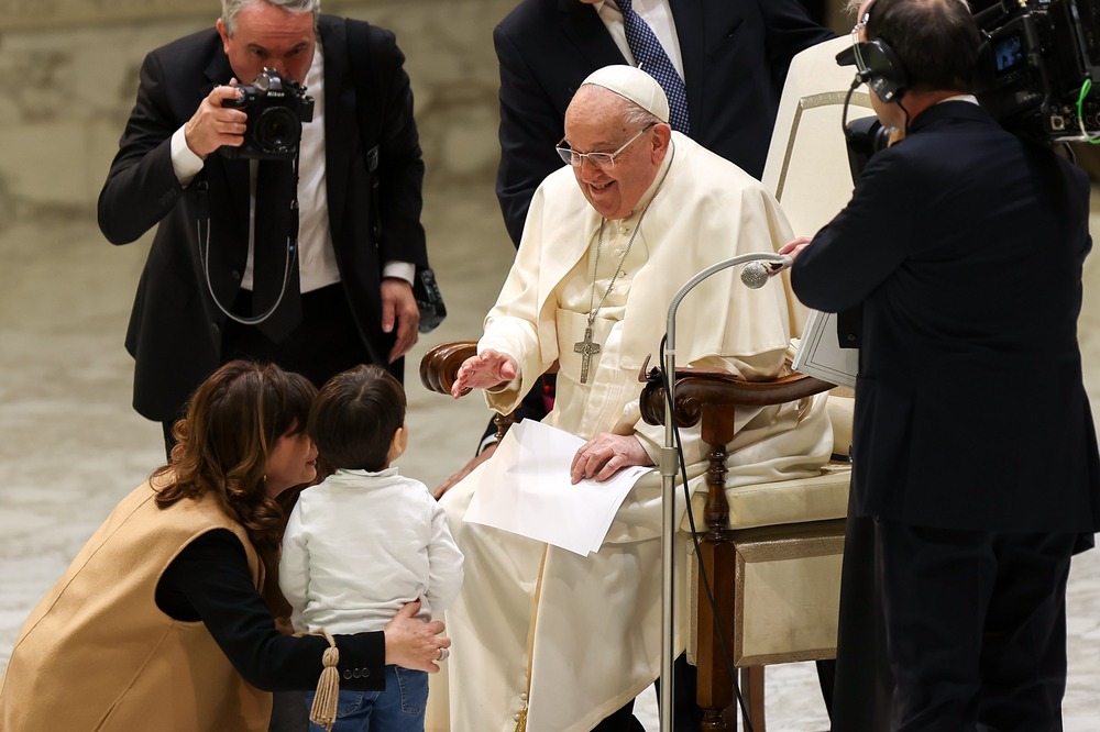 Francis seated smiles and talks to small child.