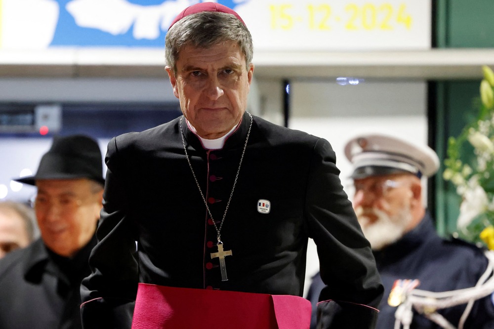 The archbishop seen in black cassock trimmed with purple walking with solemnity.