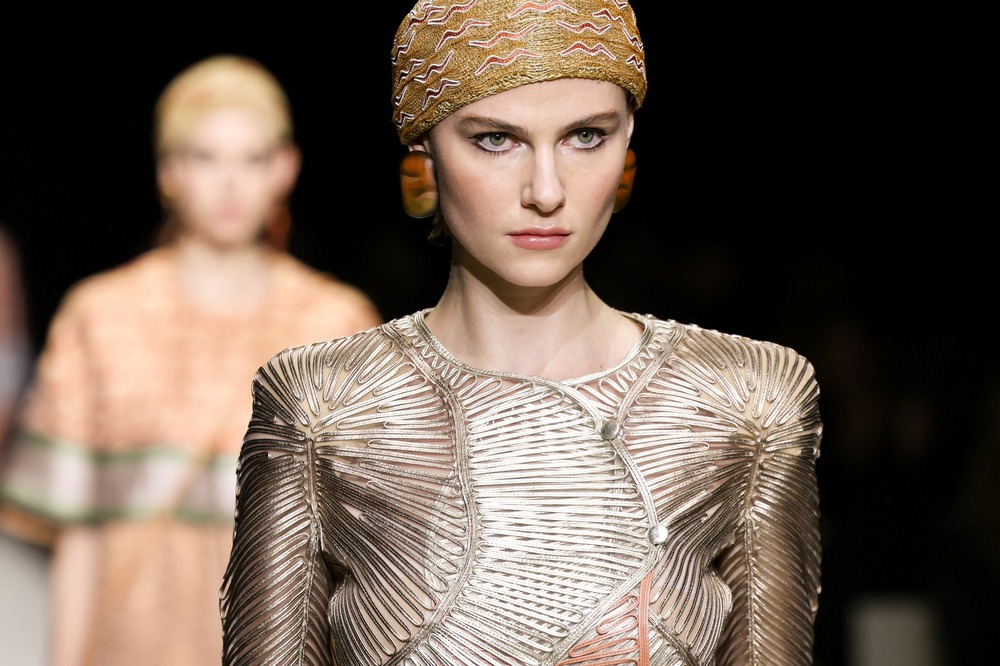 Model on runway foregrounded staring off camera, model behind seen out of focus. Garments metallic and earth-toned.