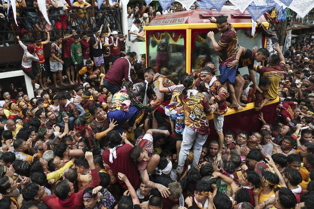 Large glass carriage contains life-size statue; it is surrounded my mammoth crowd who cling and pull to the carriage.