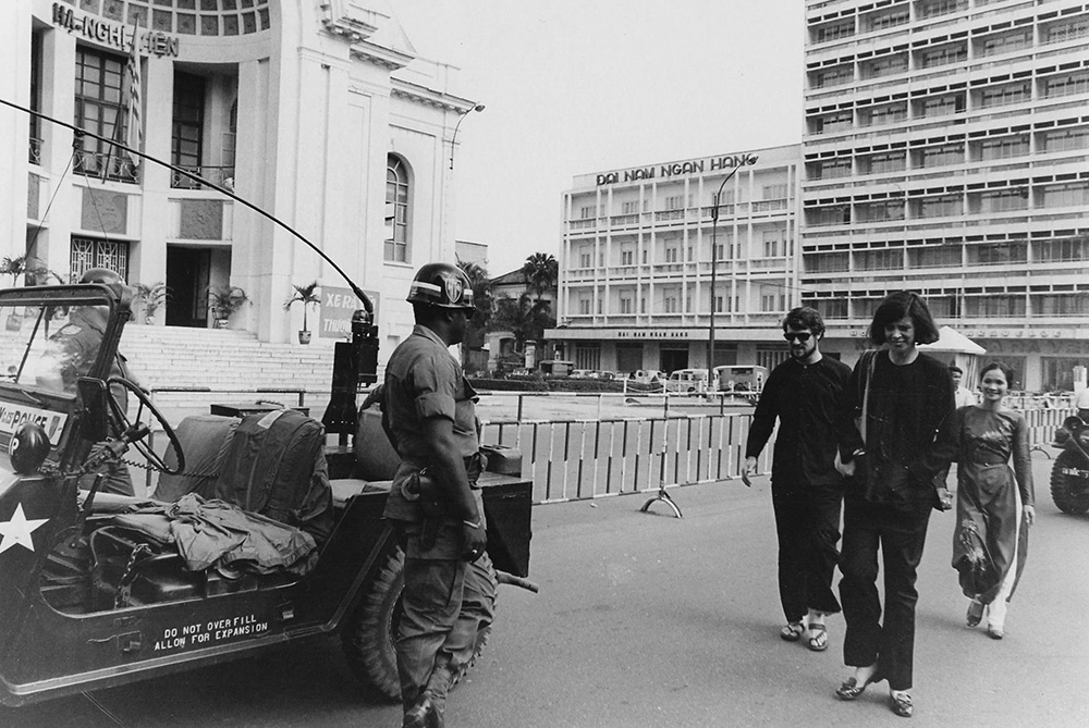 Tom Fox; Gloria Emerson, a correspondent for The New York Times; and Hoa Fox walk through downtown Saigon circa 1972, dressed in traditional Vietnamese farmer attire — which were often mistakenly identified as Viet Cong uniforms — while wary U.S. Military Police observe from a distance. (Courtesy of Thomas C. Fox)