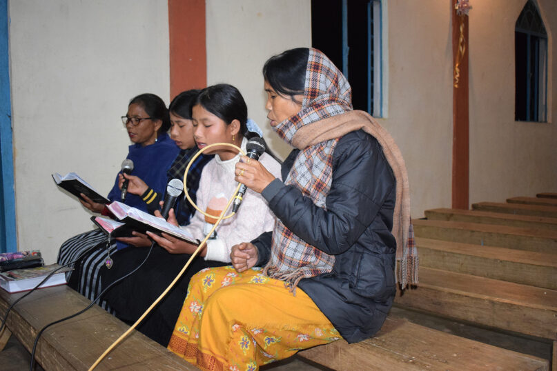 Dkhar, sits on bench in church singing into microphone.