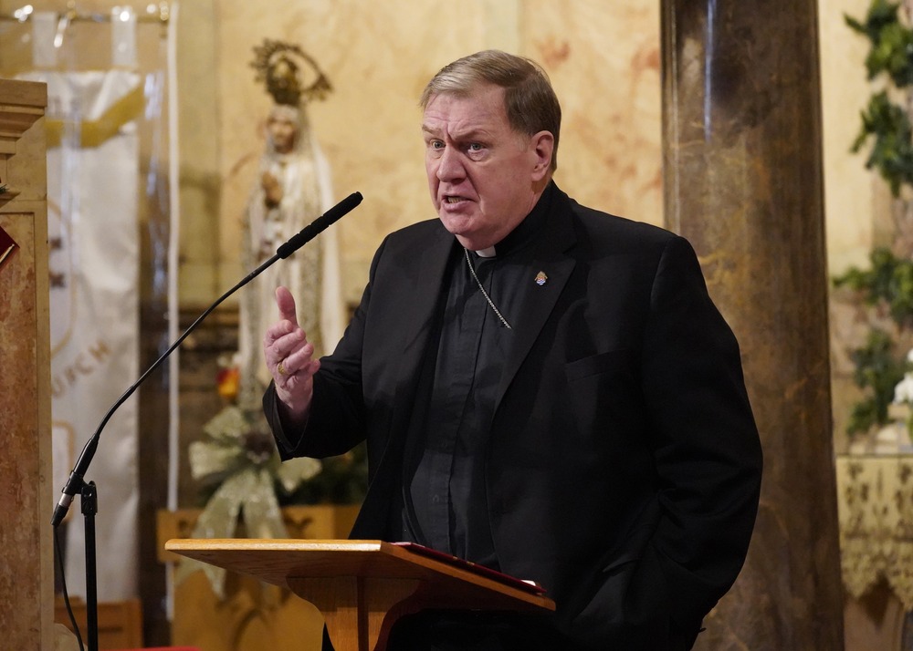 The Cardinal wears plain black clericals, stands at lectern speaking. 