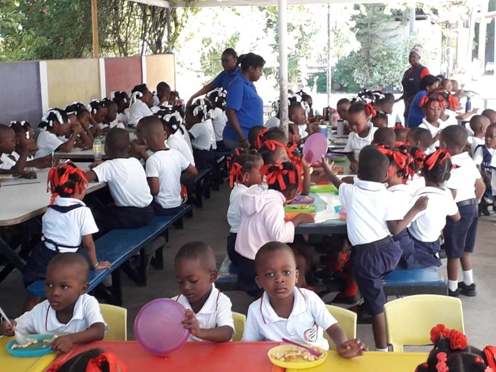 Large group of children sit along tables in covered outdoor area.