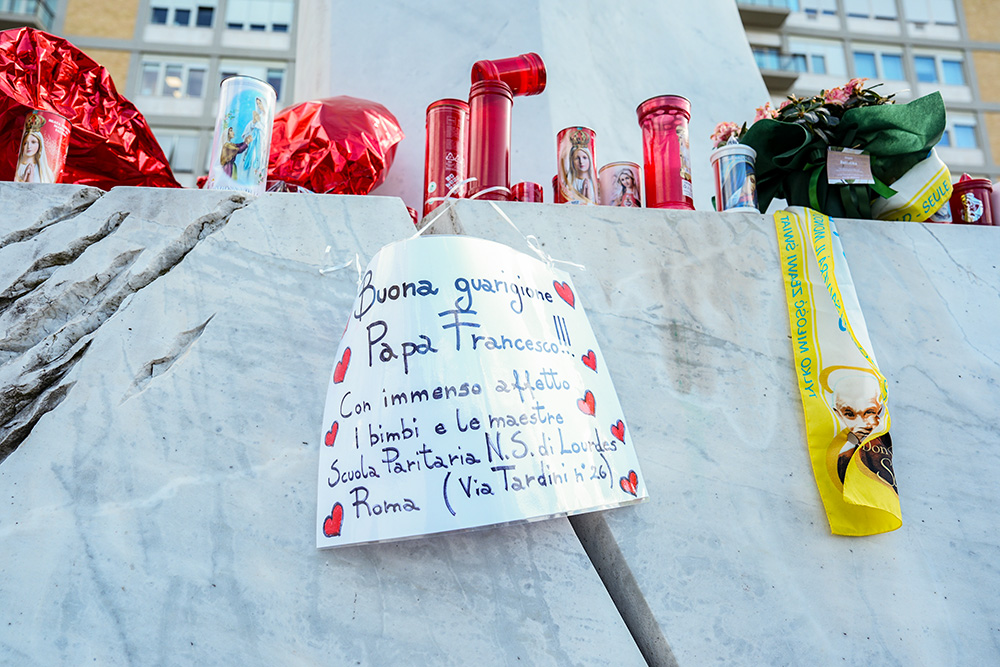 A handwritten sign with prayers for Pope Francis’ recovery is seen among candles and flowers at the base of a statue of St. John Paul II outside Rome’s Gemelli hospital Feb. 27, 2025. The pope has been hospitalized there since Feb. 14. (CNS/Lola Gomez)