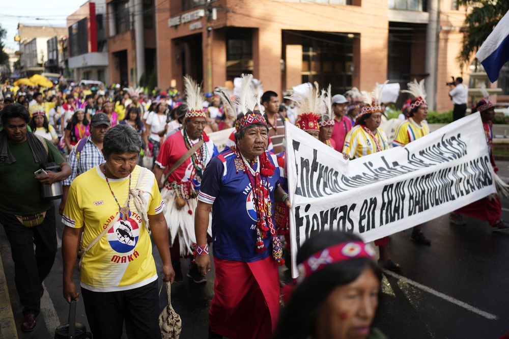 People in traditional garments march through streets behind banner.