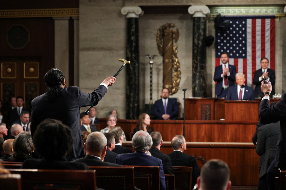 Green shown from behind shaking cane while Trump, seated, smiles smugly. 