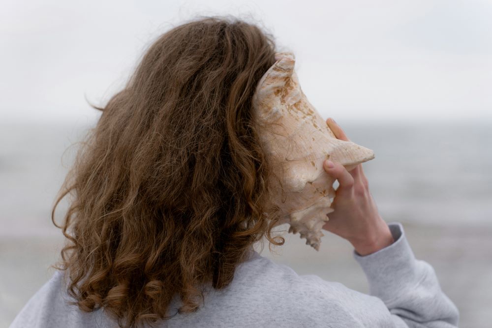 Woman faces ocean and holds shell to her ear.