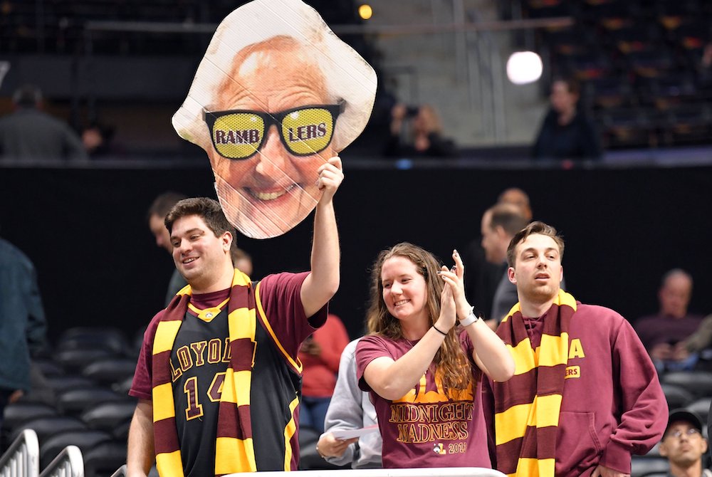 Three Loyola University Chicago students dressed in team colors, maroon and yellow, hold up a cardboard poster of Sr. Jean Dolores Schmidt's head