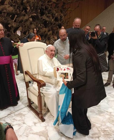 Pope seated, shakes hands and smiles with person pictured from back. 