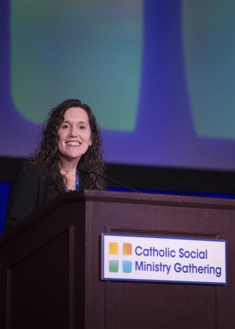 Rauh stands at a podium in front of a screen, smiling. The podium bears a sign reading "Catholic Social Ministry Gathering."