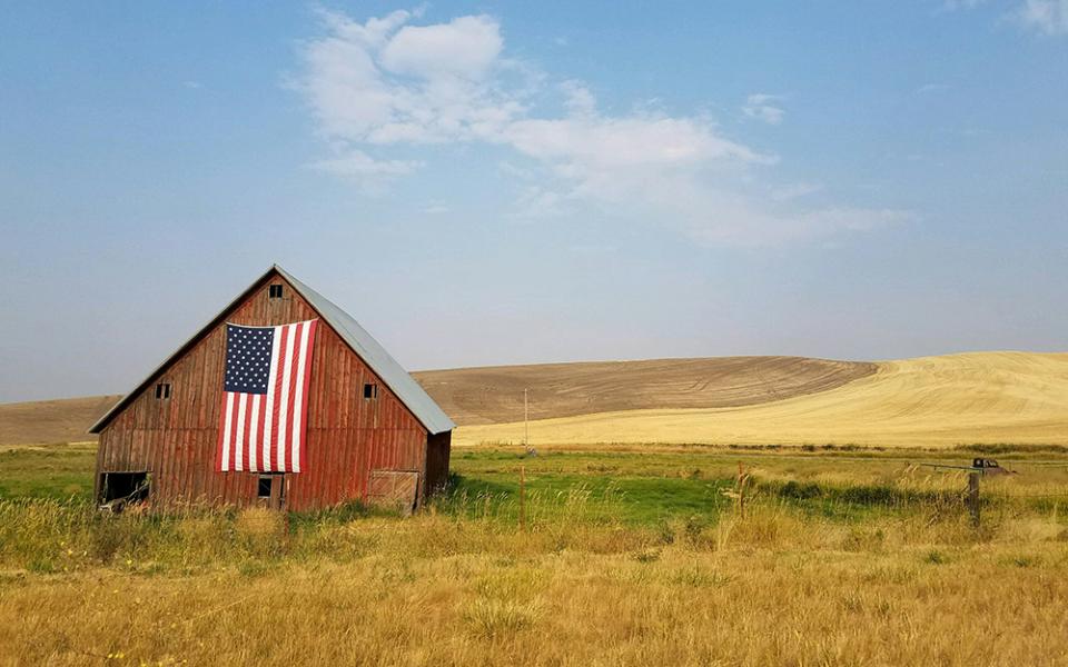 A red barn sits in the middle of a field with an American flag hanging on its side (Unsplash/specphotops)