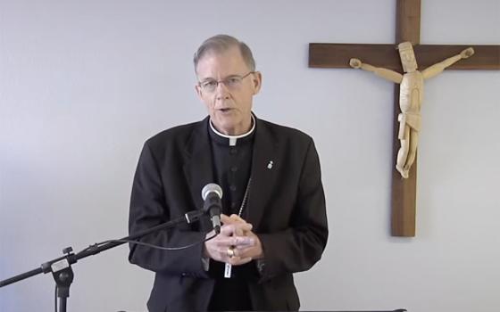 A older, white cleric stands at a lectern with a microphone in front of a crucifix