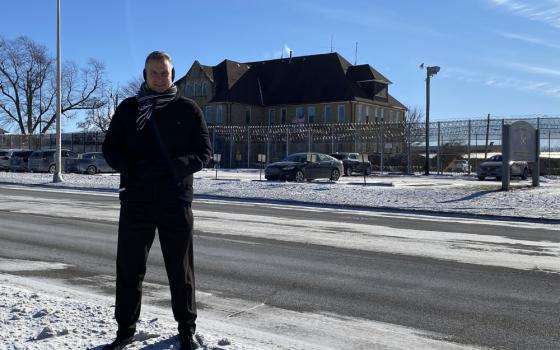 A white man wearing a black coat stands in the snow on the side of a road in front of a building