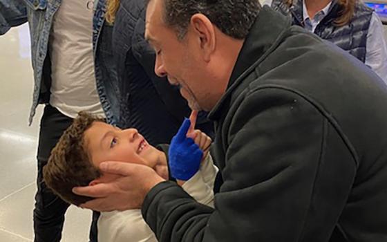 A man in  a black jacket grips a little boy's face with both hands as the little boy beams up at him