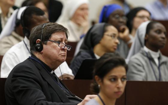 A man in a black cassock wears headphones and sits among women religious and other people