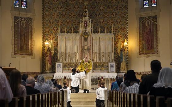 A priest with his back to the pews full of people, where some women wear lace veils, lifts a Communion host high