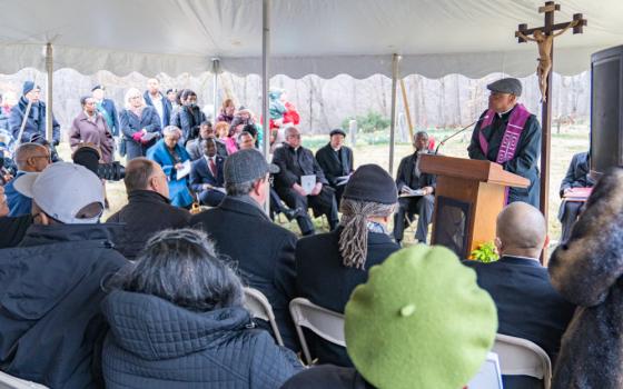 A Black man wearing a tweed cap, a black cassock, and a purple stole speaks to a large group of seated people under a tent. A crucifix is in the background