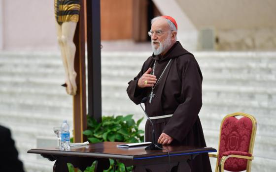 A older white man with a beard and glasses wears a brown tunic and red zucchetto and stands by a crucifix