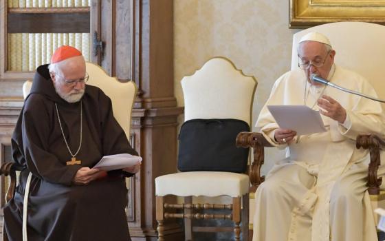 Pope Francis speaks into a microphone next to a man in a red zucchetto and a brown tunic