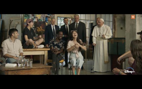 Casually dressed young people laugh as Pope Francis, standing with a cane, gestures animatedly