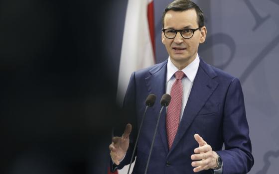 A skinny white man with glasses and a suit makes a gesture with his hands and speaks into a microphone