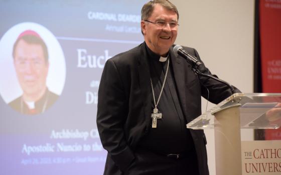 A white man in a black cassock wearing a silver pectoral cross speaks at a clear lecturn into a microphone