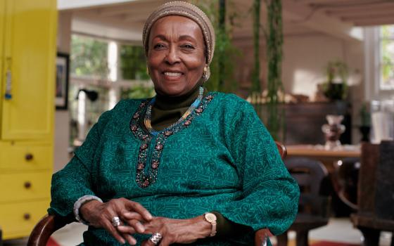 An older well-dressed Black woman wearing a head covering sits in a chair and smiles at the camera