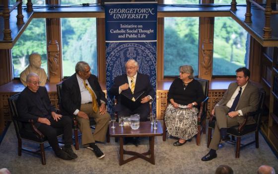 Five adults sit in front of a panel of windows. Panelists are close together and dressed up