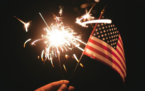 A hand holding a lit sparkler and a small American flag (Unsplash/Stephanie McCabe)