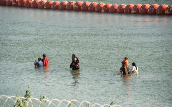 People stand in the middle of a river. Some have others on their shoulders. One part of the river is blocked by large orange buoys spaced so close as to be a wall and the other side is blocked by concertina wire.