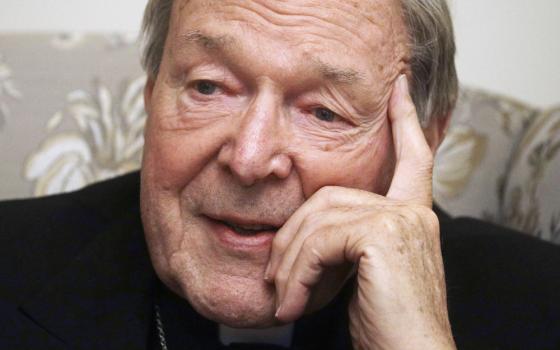 An older white man wearing a clerical collar has his pointer finger on his forehead as his other fingers rest on his cheek