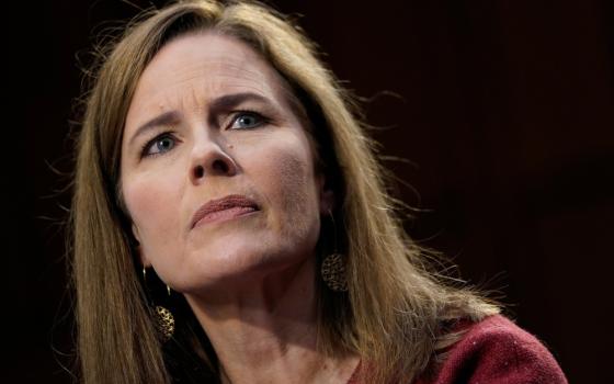 A white woman with shoulder length brown hair wearing a red sweater, dangly earrings and light makeup stares off camera 