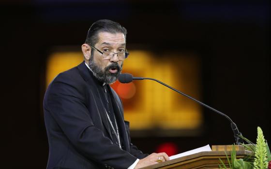 A light-skinned man with a beard and glasses and black hair speaks into a microphone at a podium