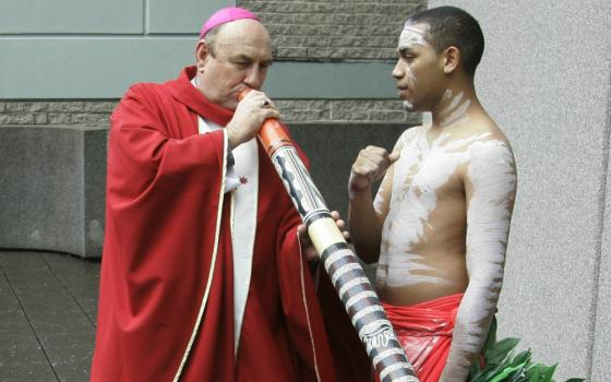 A white man wearing a pink zucchetto and red vestments blows into a digeridoo while standing next to a dark-skinned youth wearing white body paint.