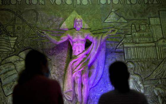 The shadows of two people are visible before a stone statue of Jesus that stands in front of the words, "Estoy con vosotros todos los dias."